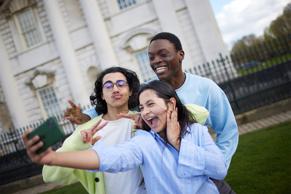 A group of students taking a selfie with one of the Greenwich Campus buildings in the background