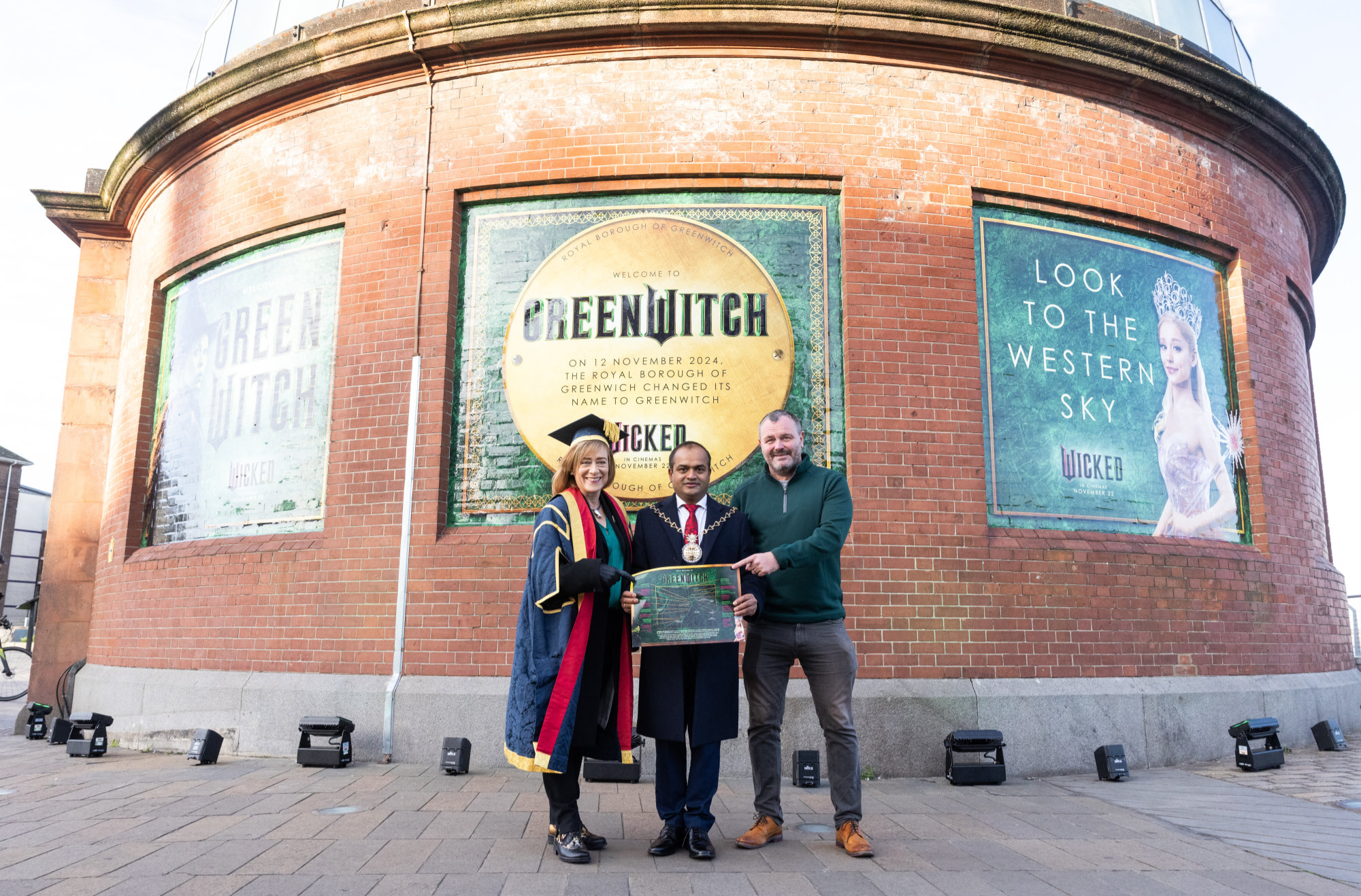 Three people holding up a map of the GreenWitch trail in front of the entrance to the greenwich foot tunnel